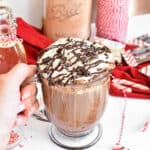 Hand holding a clear mug of peppermint mocha topped with whipped cream, chopped peppermint bark and a large drizzle of chocolate sauce