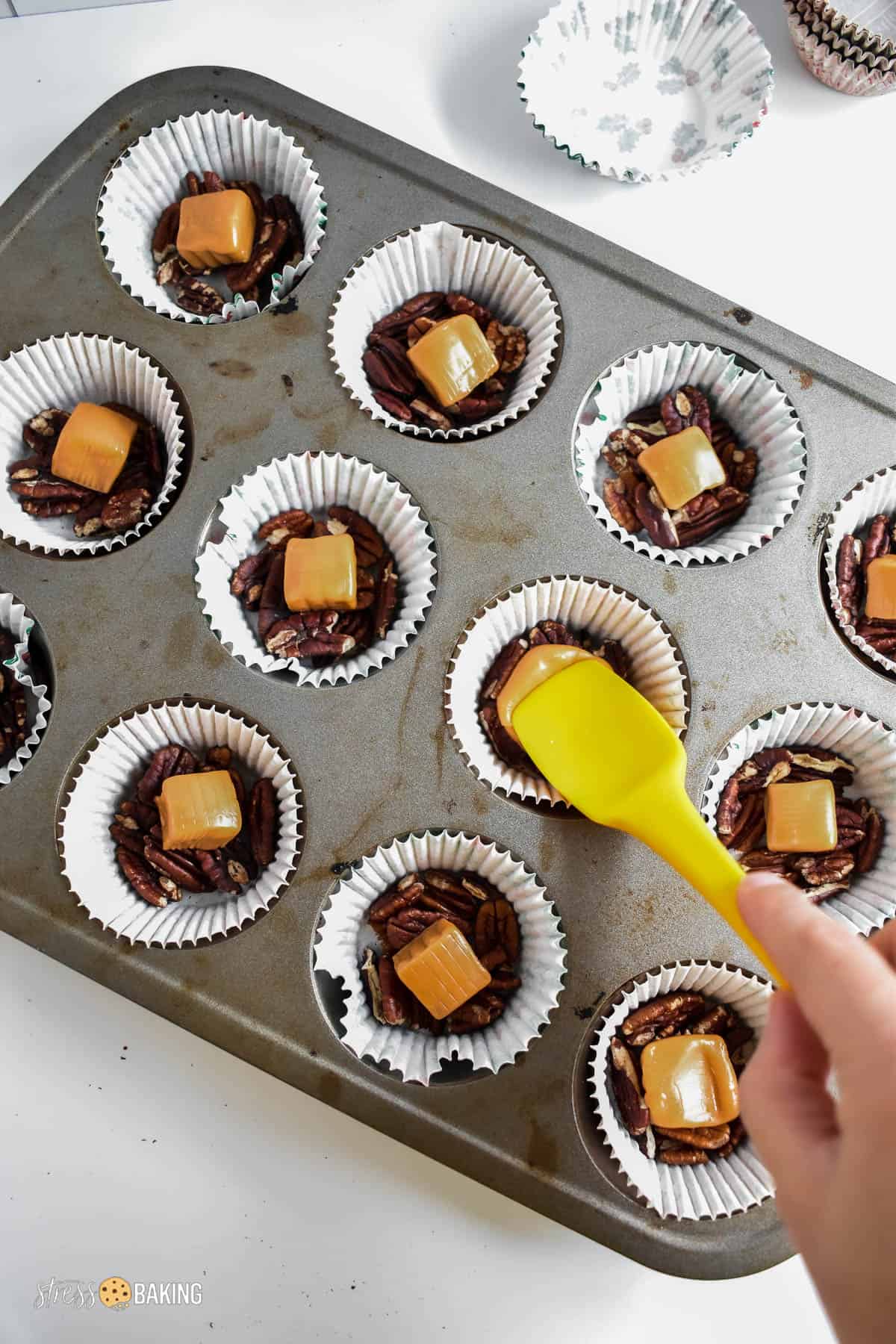 Muffin pin filled with cupcake liners filled with pecan pieces and warm caramel squares being pressed down with a yellow spatula