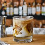 White Russian cocktail being stirred to show swirls of cream and coffee liqueur in a clear old fashioned rocks glass on a wood cutting board