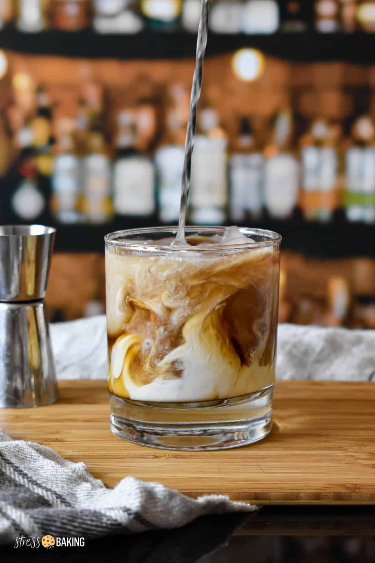 White Russian cocktail being stirred to show swirls of cream and coffee liqueur in a clear old fashioned rocks glass on a wood cutting board