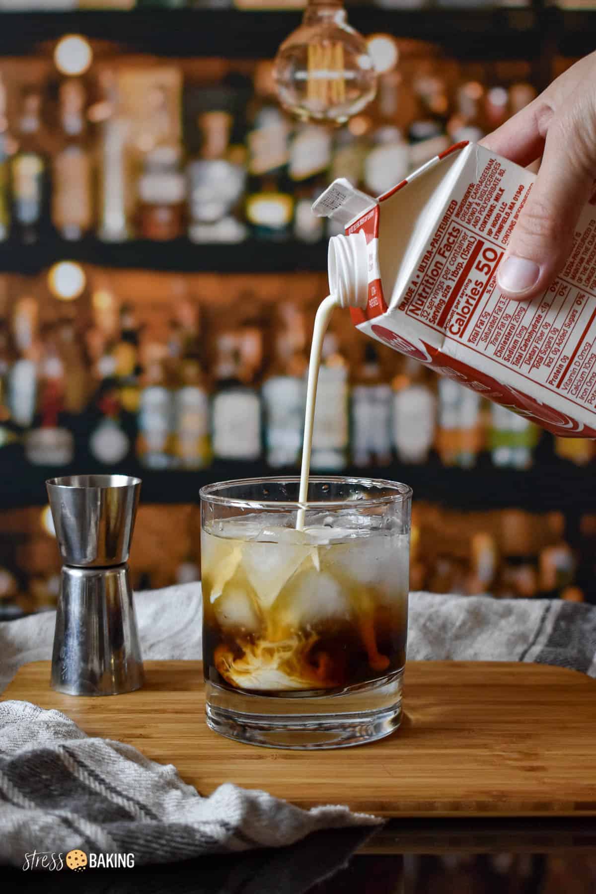 Cream being poured into an old fashioned rocks glass full of vodka and coffee liqueur and ice on a wood cutting board