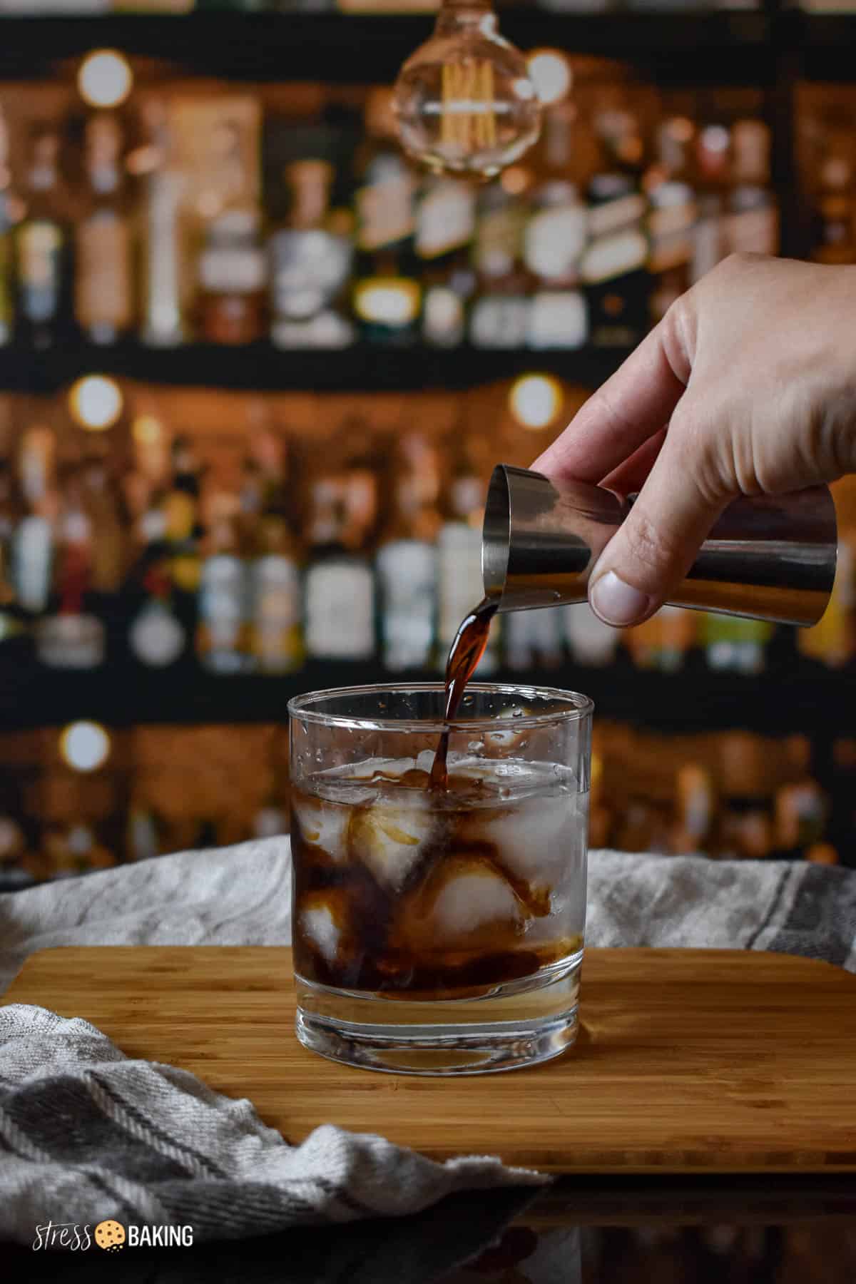Coffee liqueur being poured into an old fashioned rocks glass full of vodka and ice on a wood cutting board