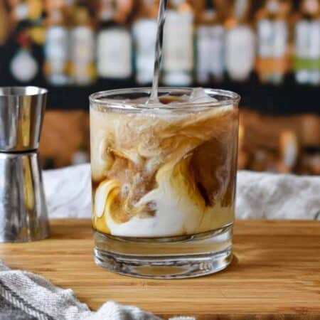 White Russian cocktail showing layers of cream and coffee liqueur in a clear old fashioned rocks glass on a wood cutting board