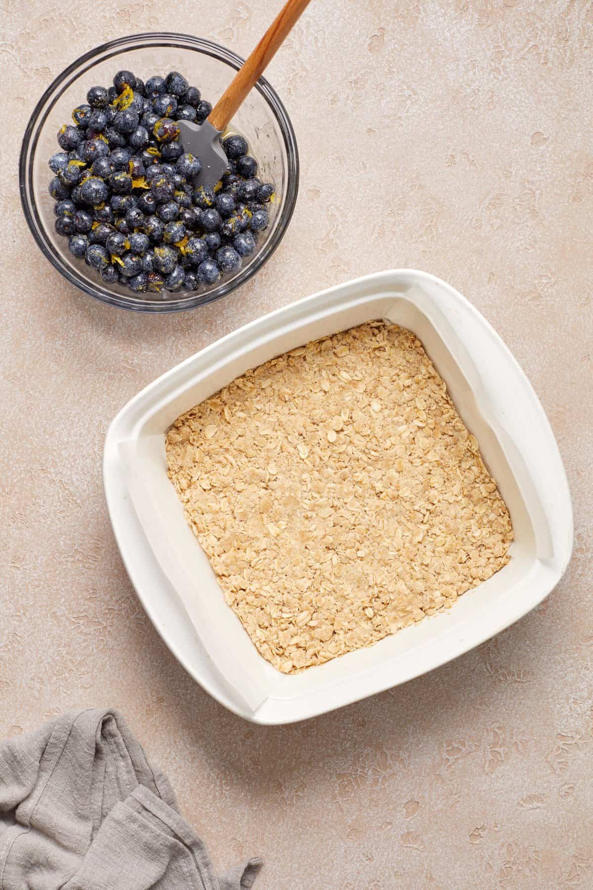 Square white baking dish filled with oatmeal mixture next to a bowl of blueberries and lemon zest