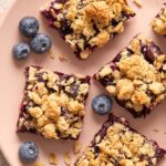 Golden oatmeal topping on slices of purple blueberry bars on a blush colored plate