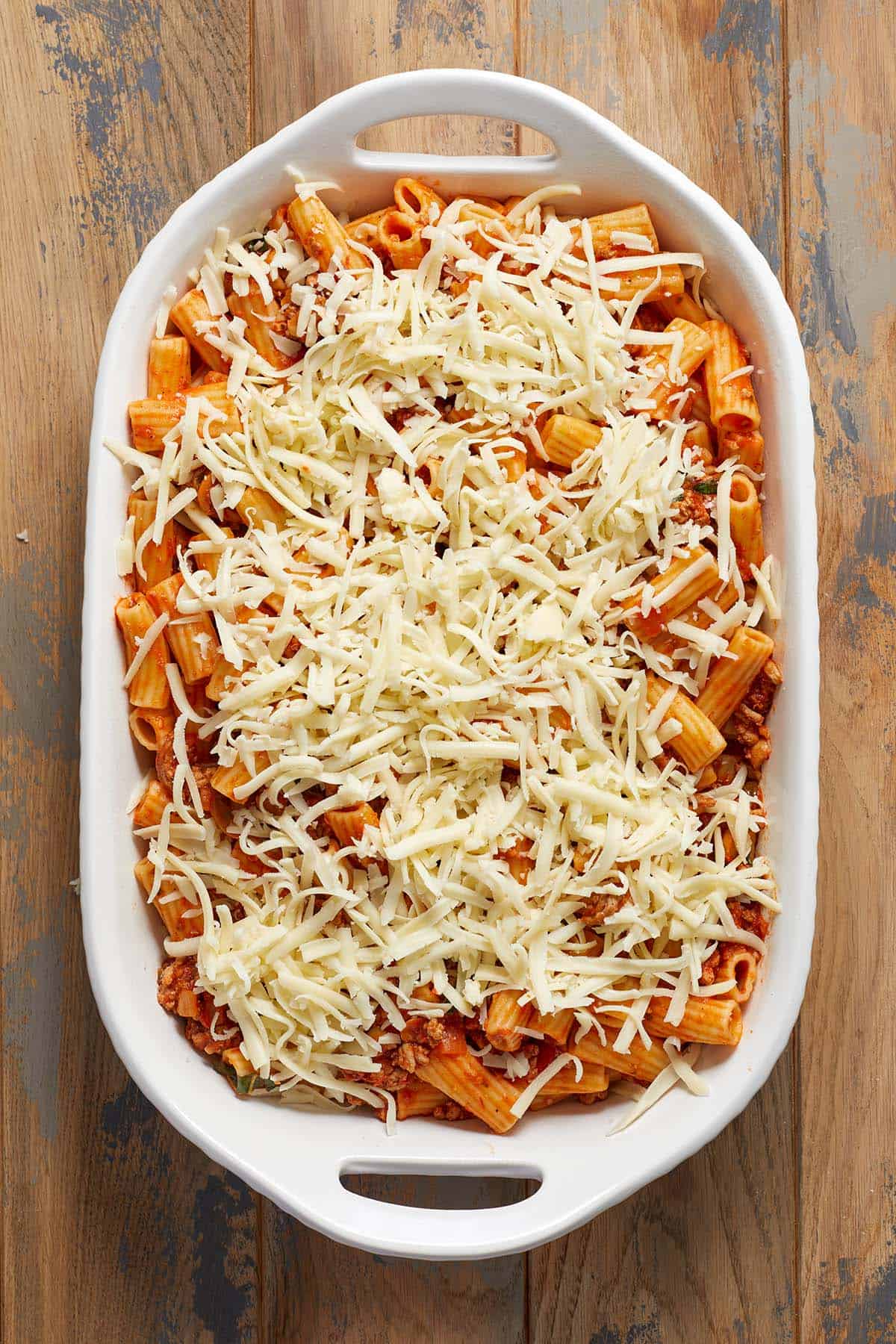 Rigatoni pasta covered in sauce and shredded cheese in a white baking dish on a wooden surface