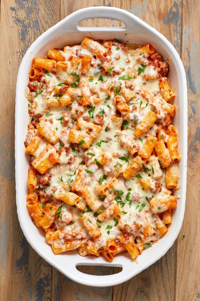 Baked rigatoni topped with melted cheese and parsley in a white baking dish on a wooden surface