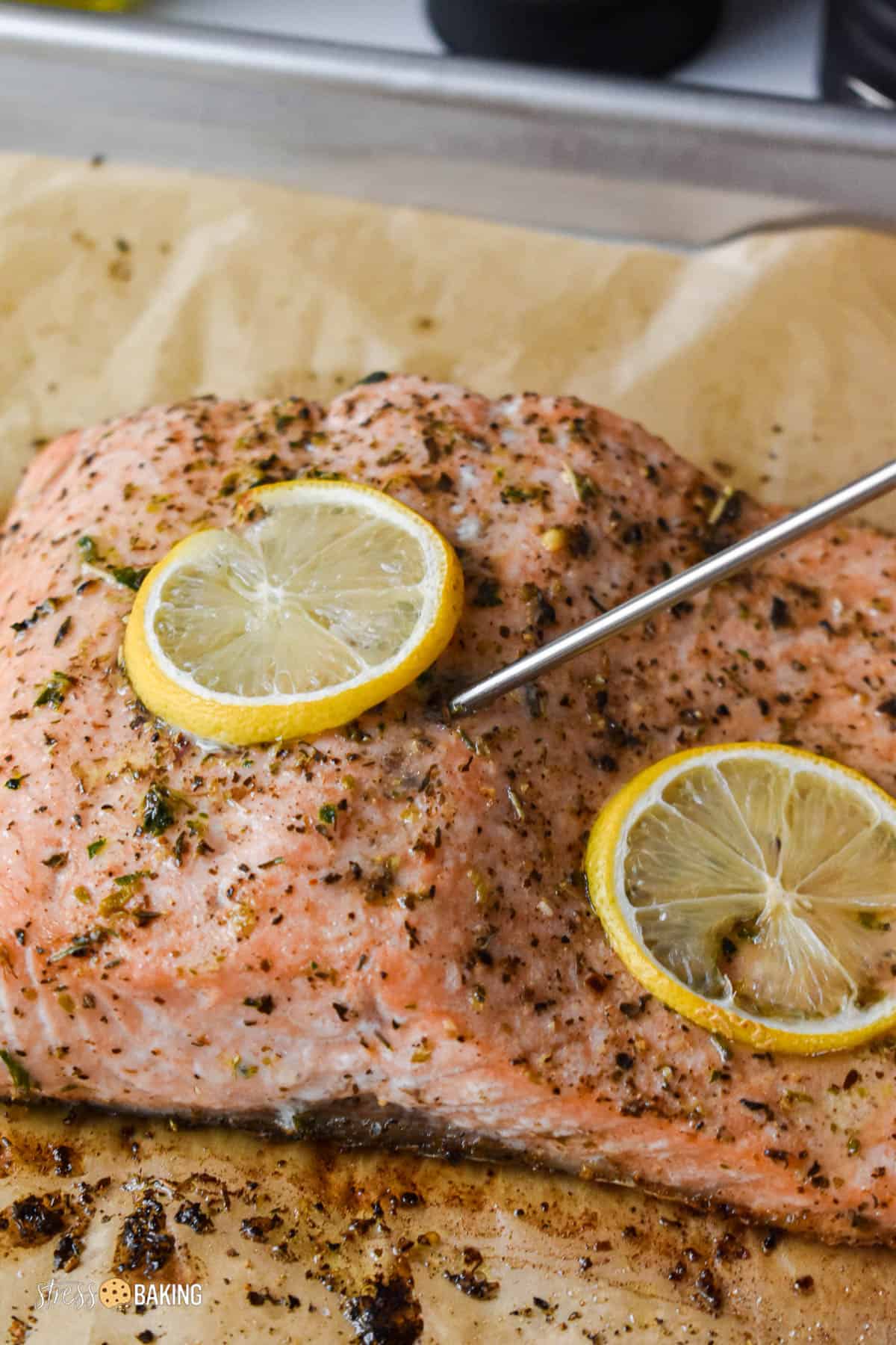 A thermometer being stuck in a piece of baked salmon coated in spices and thin lemon slices on parchment paper