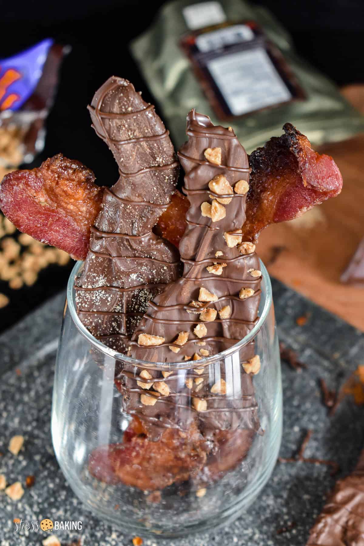 Chocolate dipped bacon strips coated in sugar and toffee pieces sitting in a clear glass