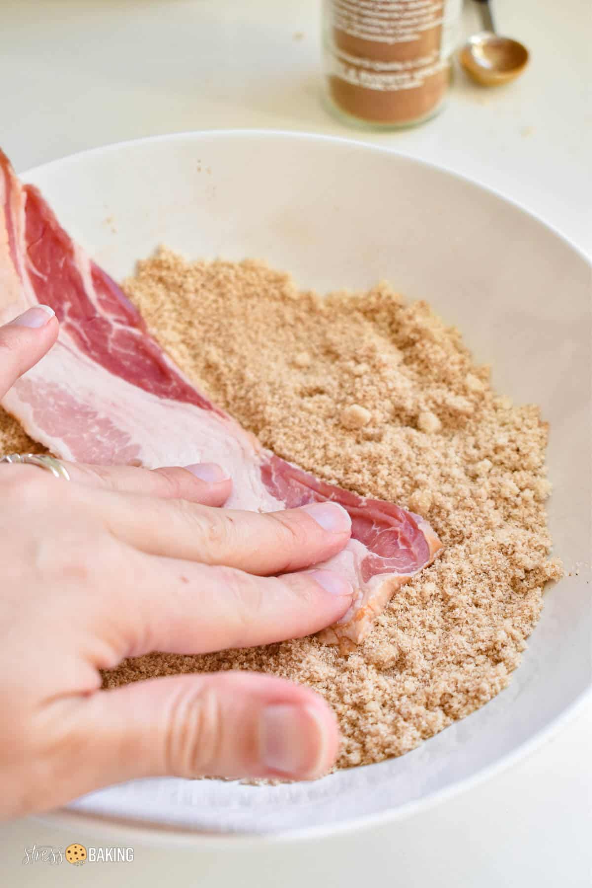 Hand pressing a piece of raw bacon into brown sugar