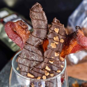 Chocolate dipped bacon strips coated in sugar and toffee pieces sitting in a clear glass