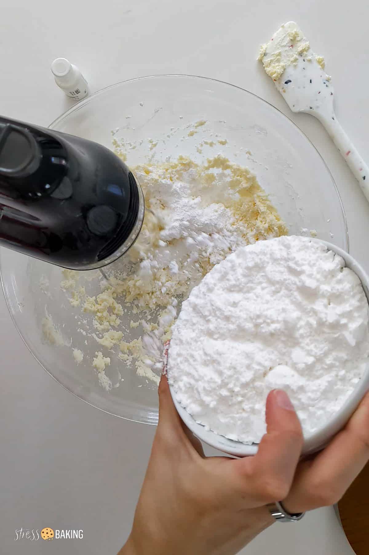 Powdered sugar being added to cream cheese mixture while a hand mixer runs on low speed