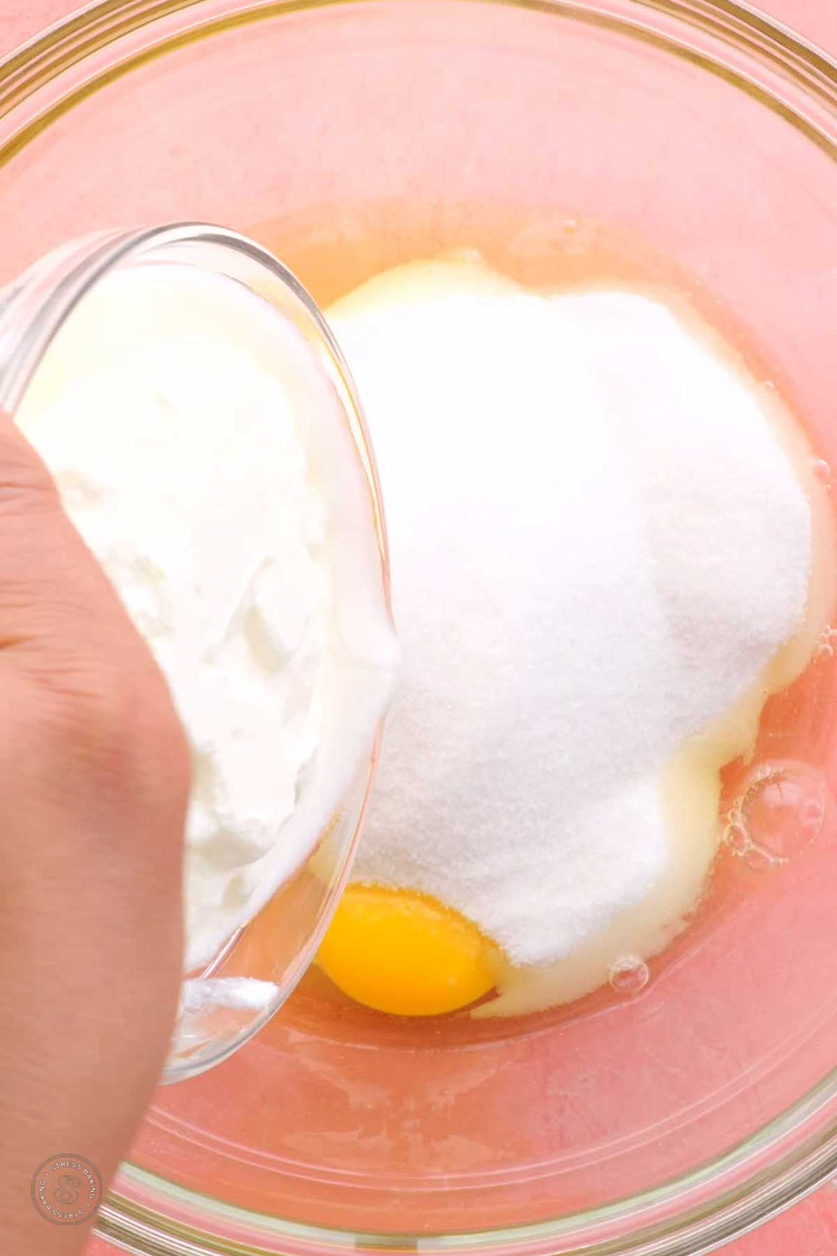 Yogurt being poured into a bowl of eggs and sugar