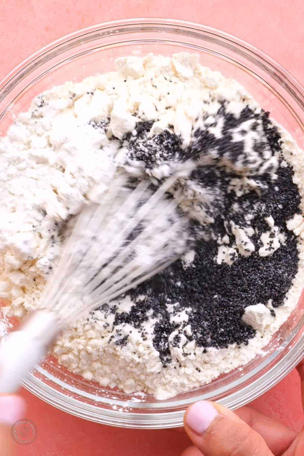 Poppy seeds being whisked together with white dry ingredients in a clear bowl