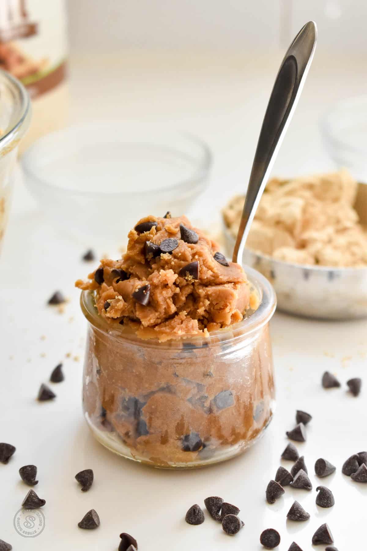 A small clear jar stuffed with peanut butter cookie dough dotted with mini chocolate chips with a small spoon stuck inside the jar