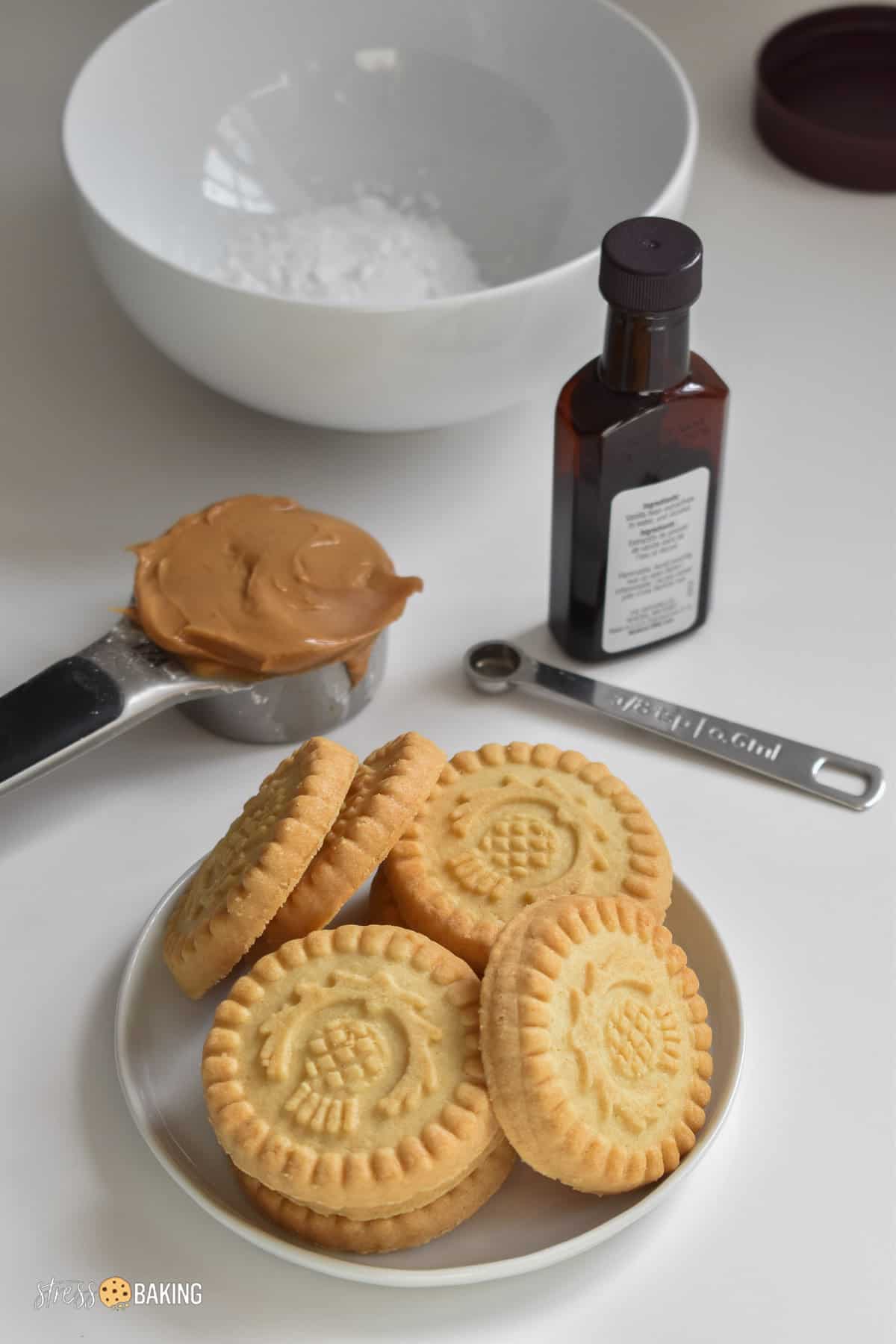 Round shortbread cookies on a white plate in front of a measuring cup of peanut butter, a jar of vanilla and a bowl of powdered sugar