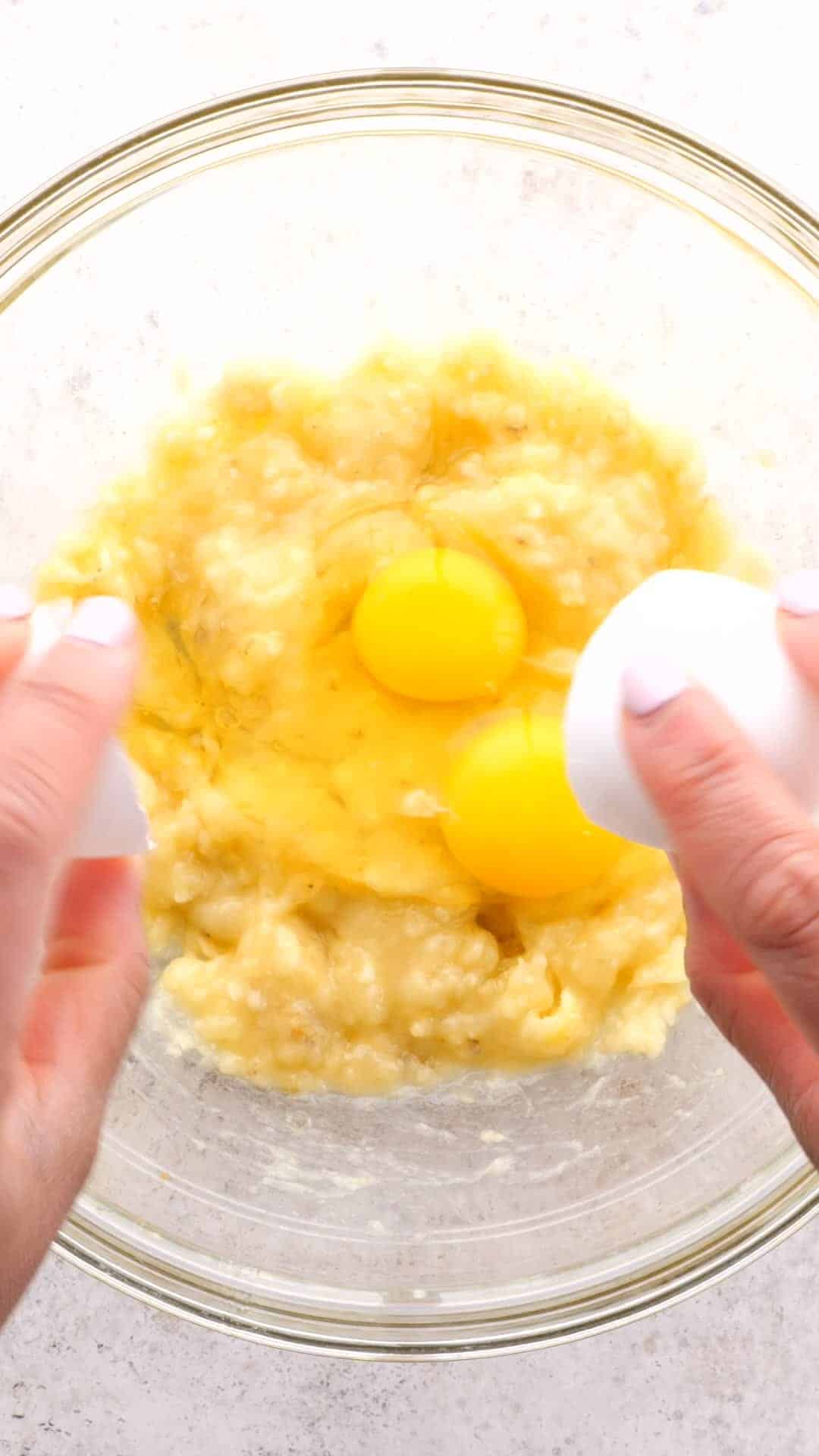 Eggs being cracked into a clear bowl of mashed bananas