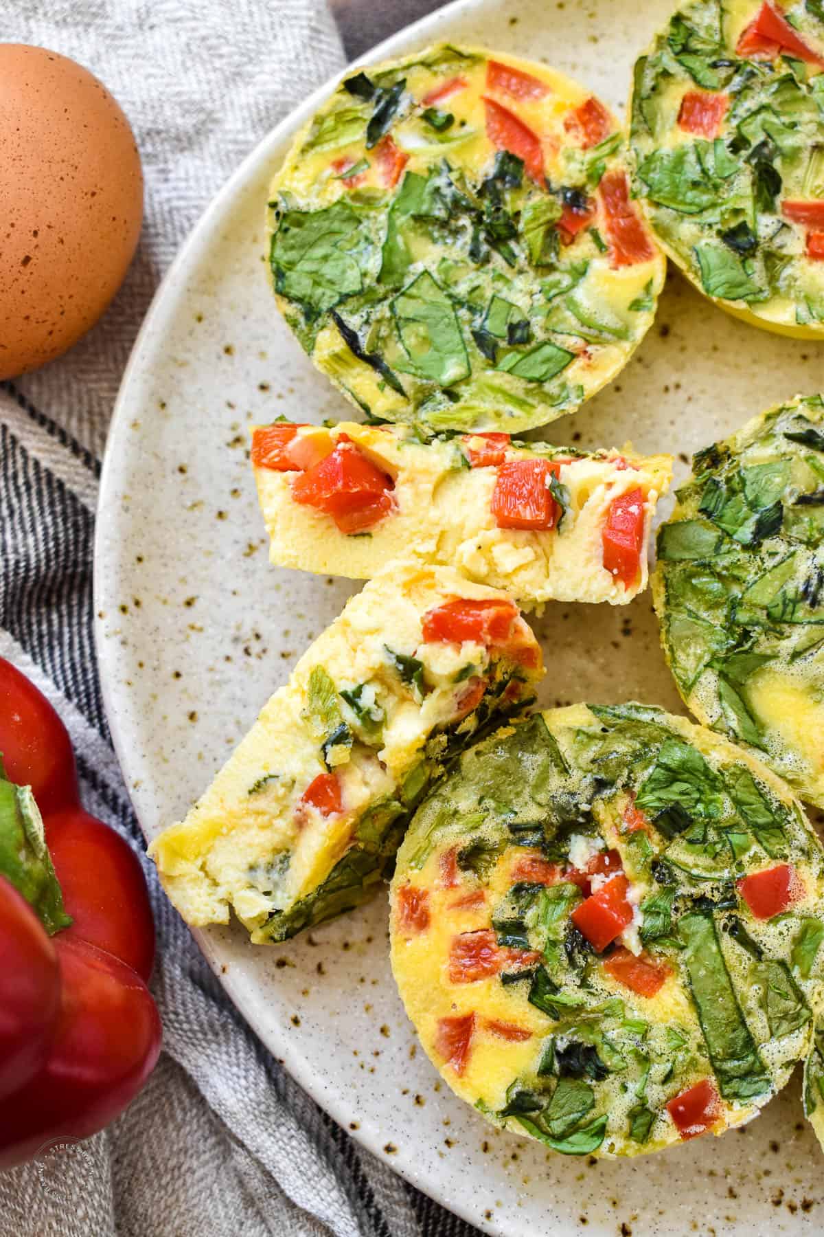 Round yellow egg bites full of spinach and diced bell pepper with one cut in half to show the velvety interior