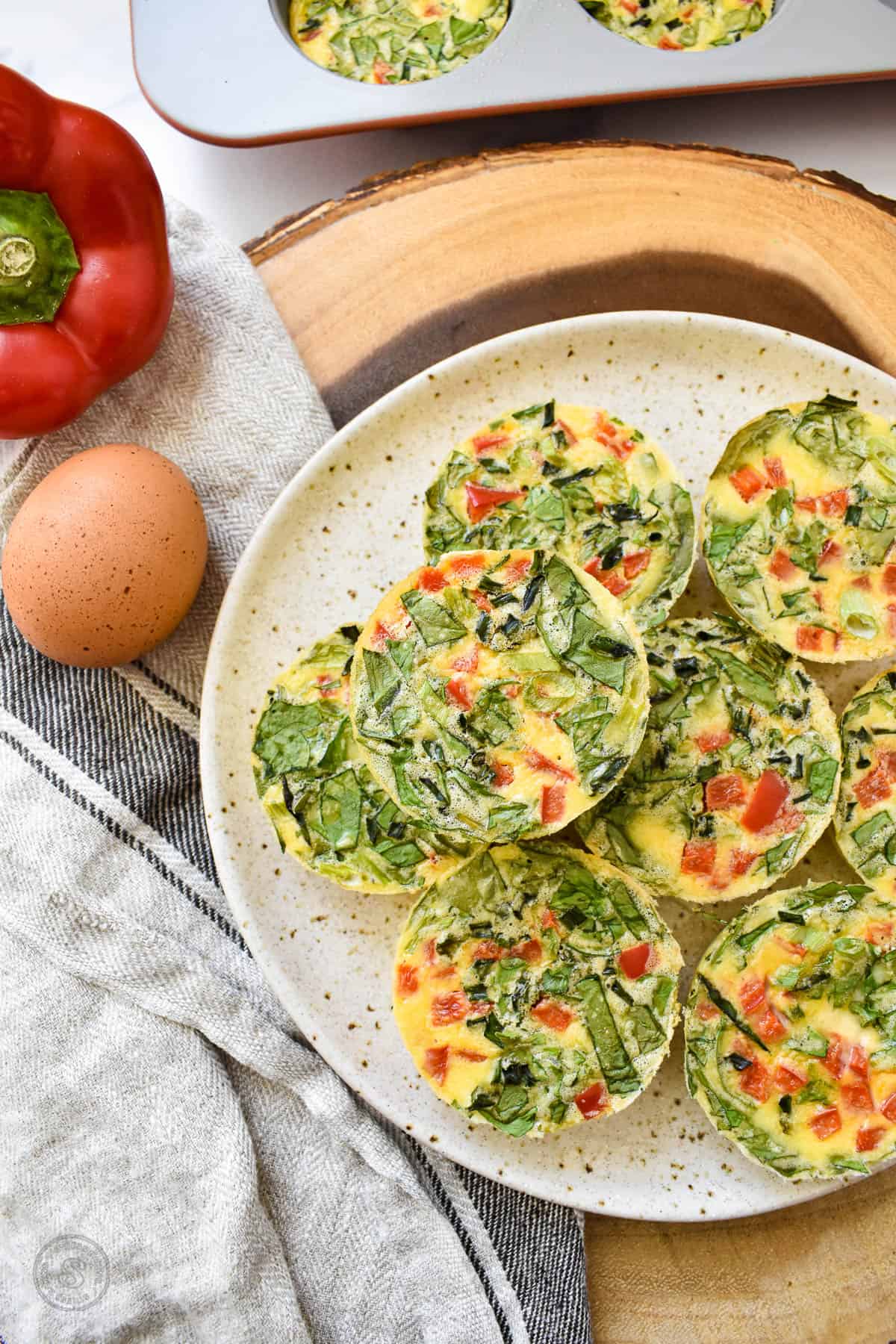 Round yellow egg bites full of spinach and diced bell pepper stacked on a tan speckled plate on a wood platter