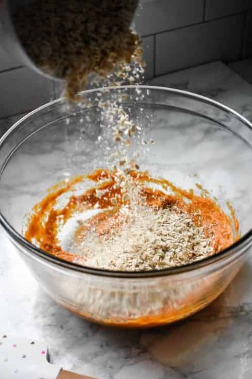 Ground oats being poured into clear mixing bowl of pumpkin puree