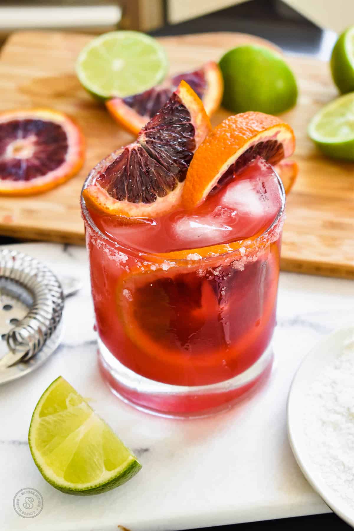 Blood orange margarita in a salt-rimmed glass topped with wedges and slices of blood orange and lime on a white marble tray