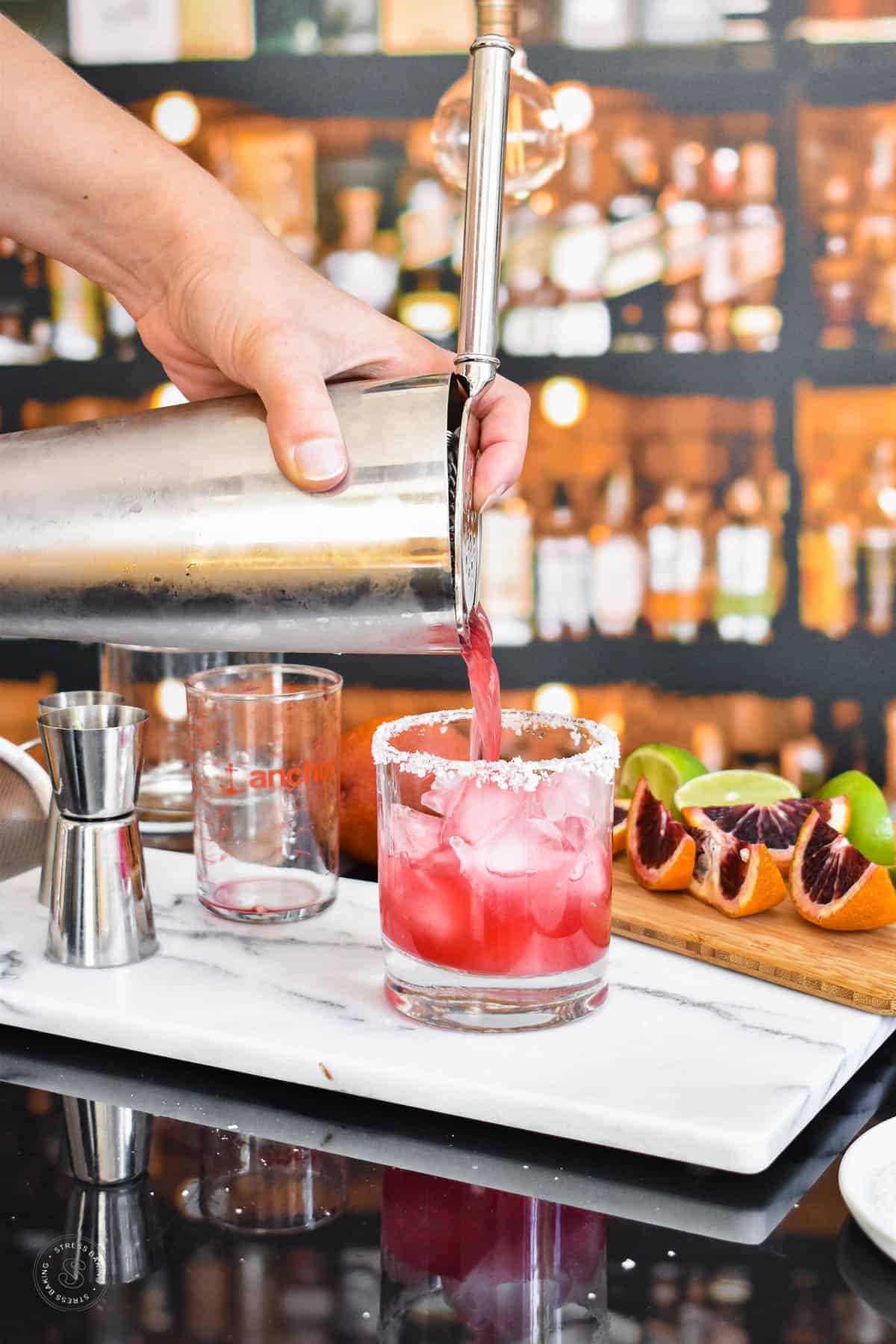Blood orange margarita being strained out of a cocktail shaker into a salt-rimmed glass a white marble tray next to a salted rimmed rocks glass