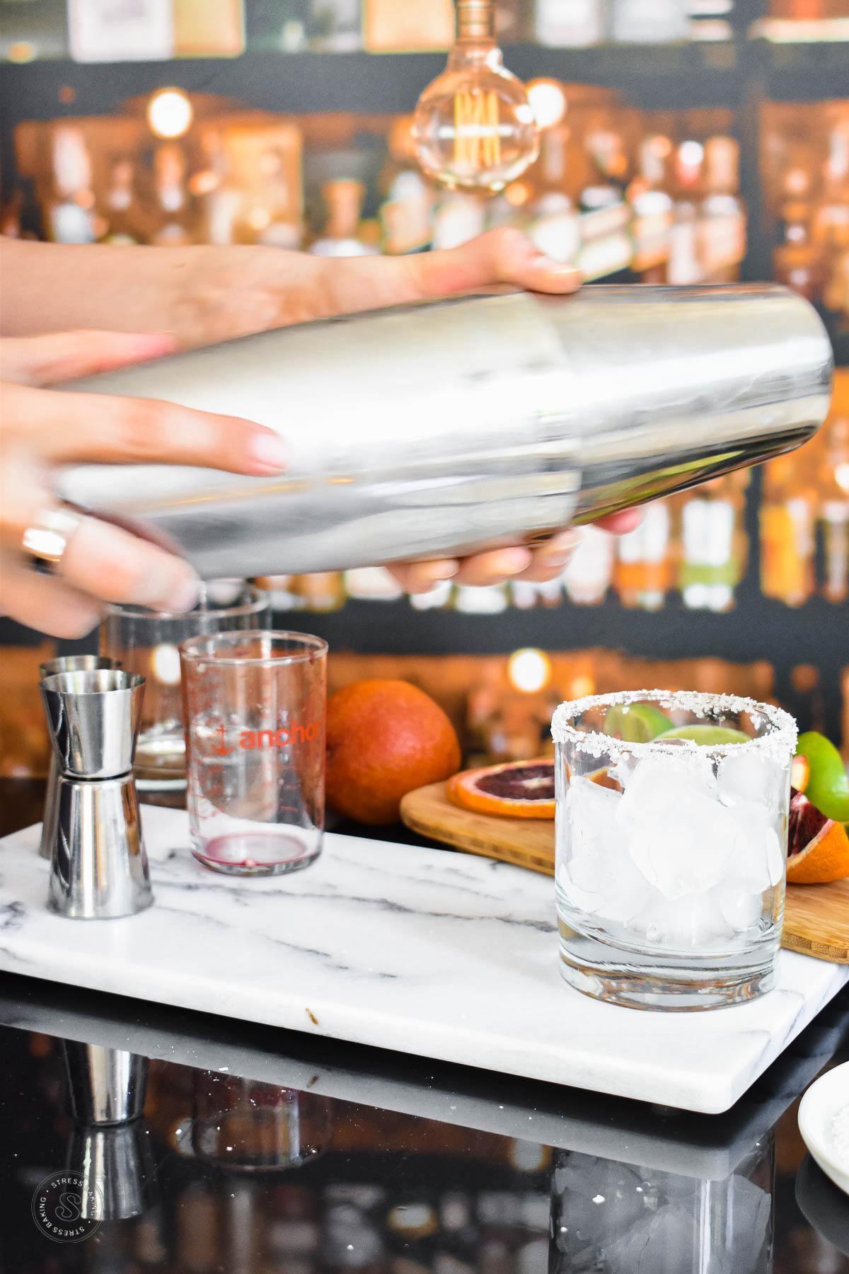 A cocktail shaker being shaken above a white marble tray next to a salted rimmed rocks glass