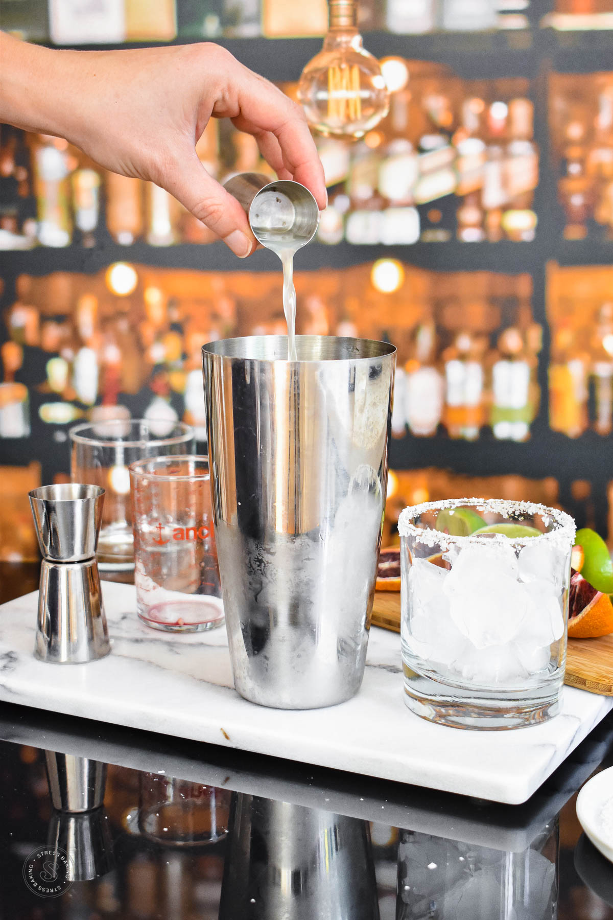 Lime juice being poured into a cocktail shaker on a white marble tray next to a salted rimmed rocks glass