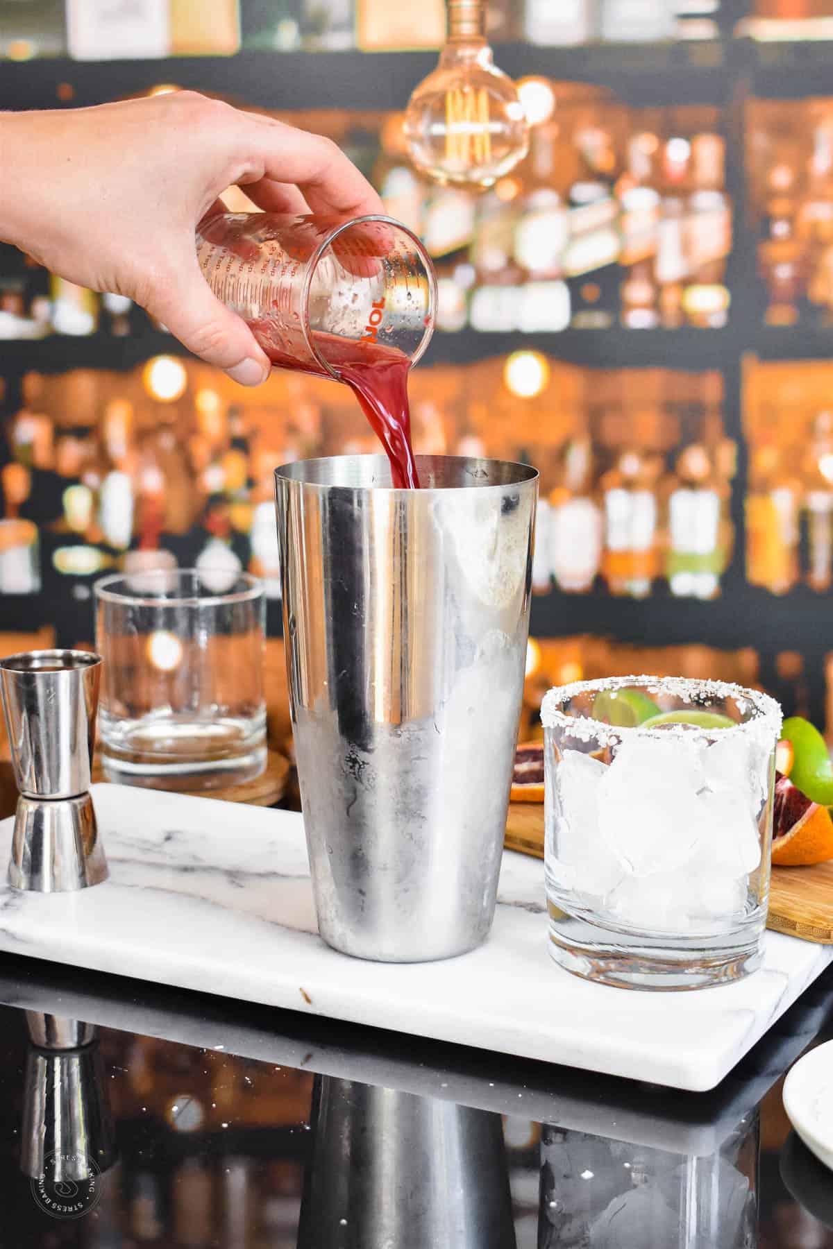 Blood orange juice being poured into a cocktail shaker on a white marble tray next to a salted rimmed rocks glass