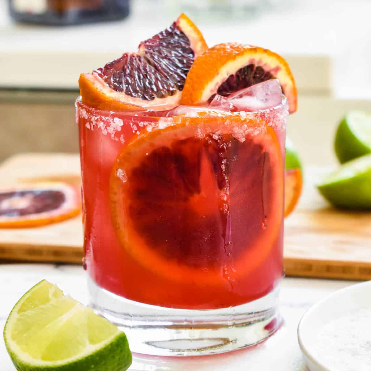 Blood orange margarita in a salt-rimmed glass topped with wedges and slices of blood orange and lime on a white marble tray