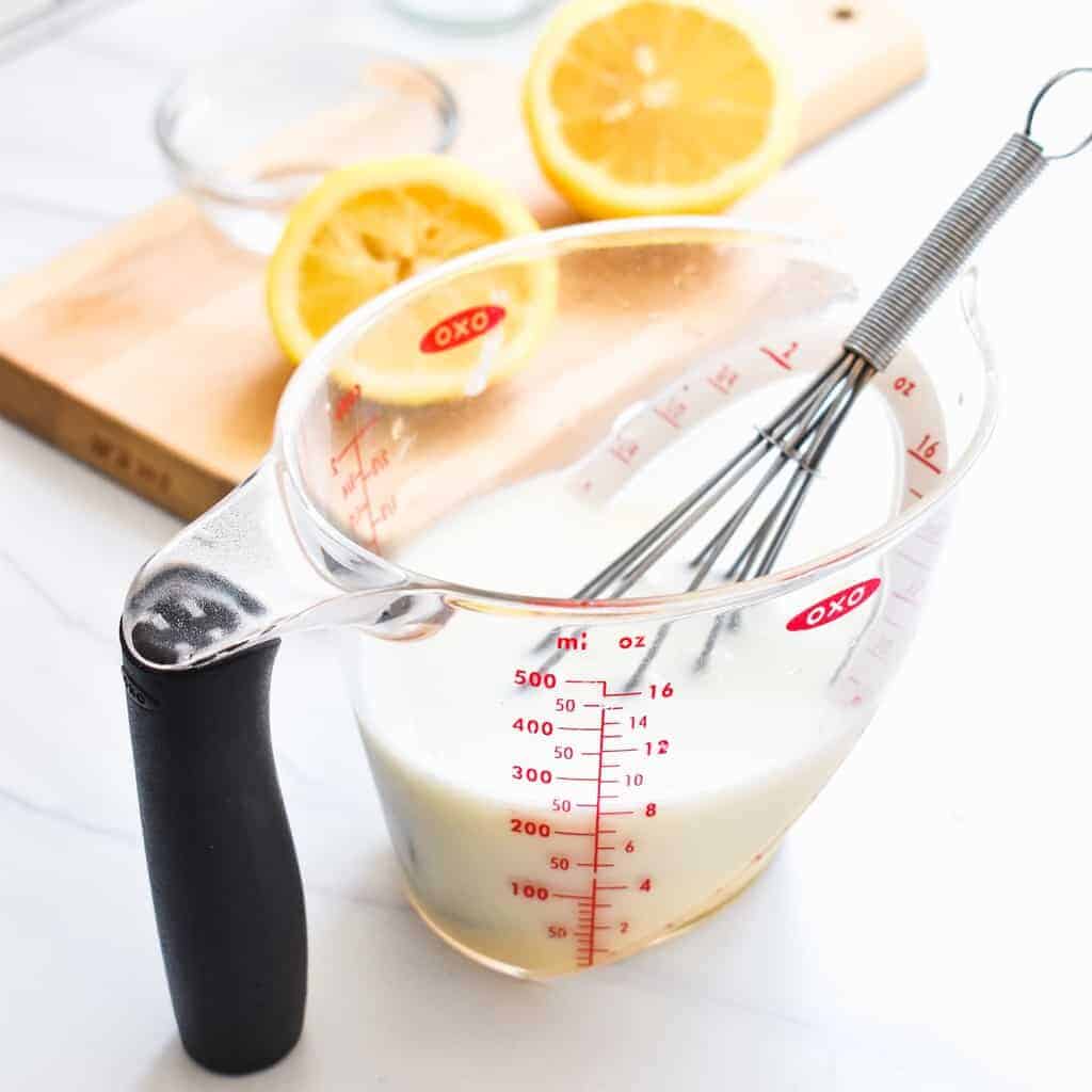 A whisk in a measuring cup of milk and lemon juice in front of a small cutting board with a sliced lemon