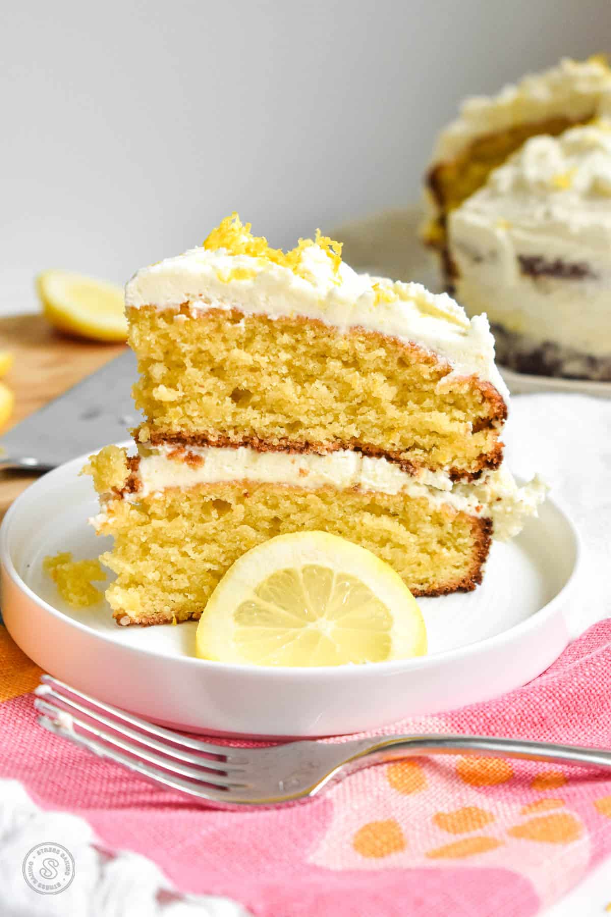 Slice of yellow lemon layer cake on a white plate next to a slice of lemon with white frosting topped with lemon zest