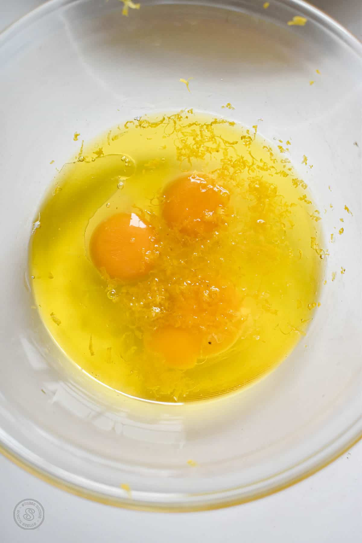 Eggs combined with oil and lemon zest in a clear mixing bowl