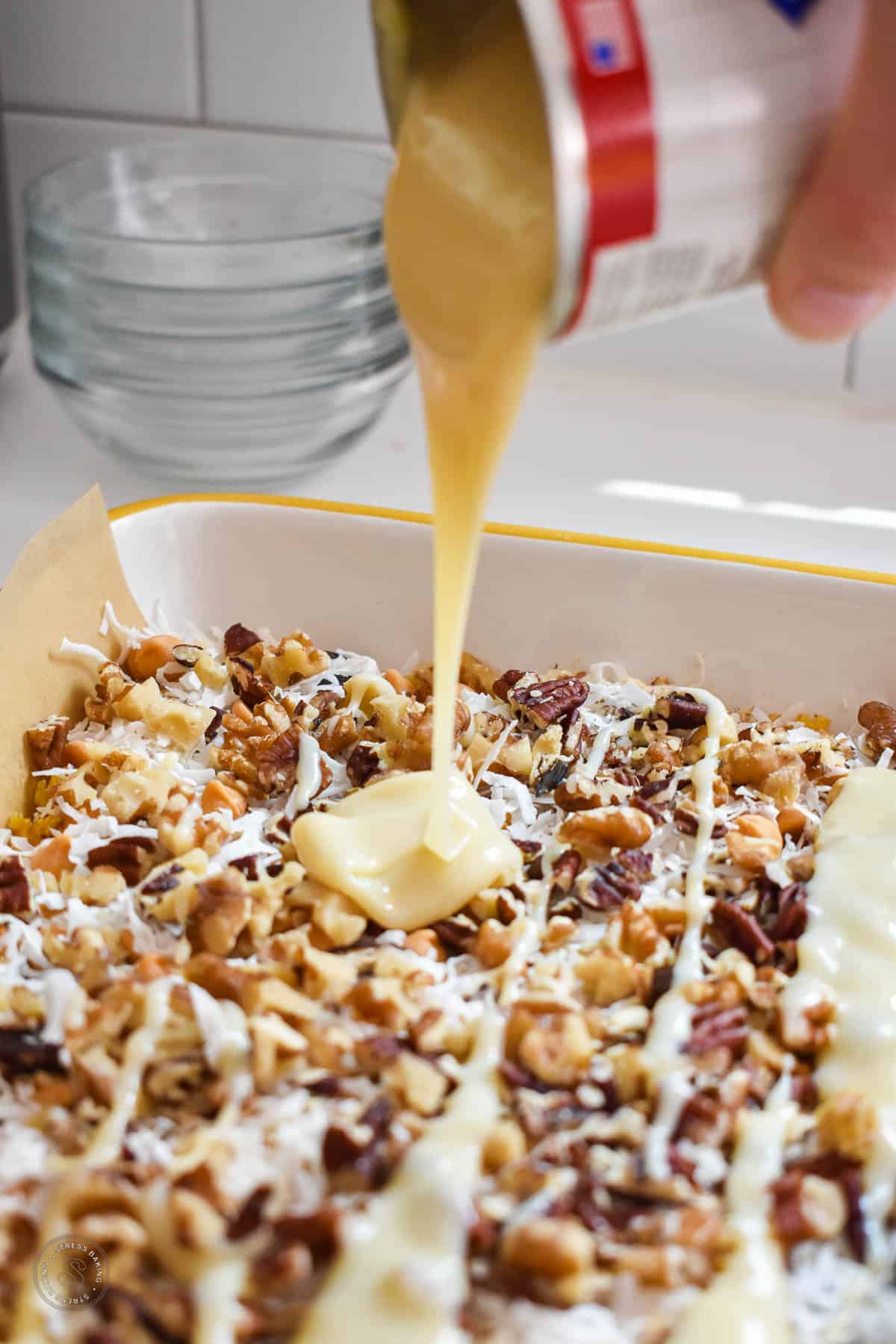 Condensed milk being poured on top of shredded coconut and chopped nuts in a baking dish