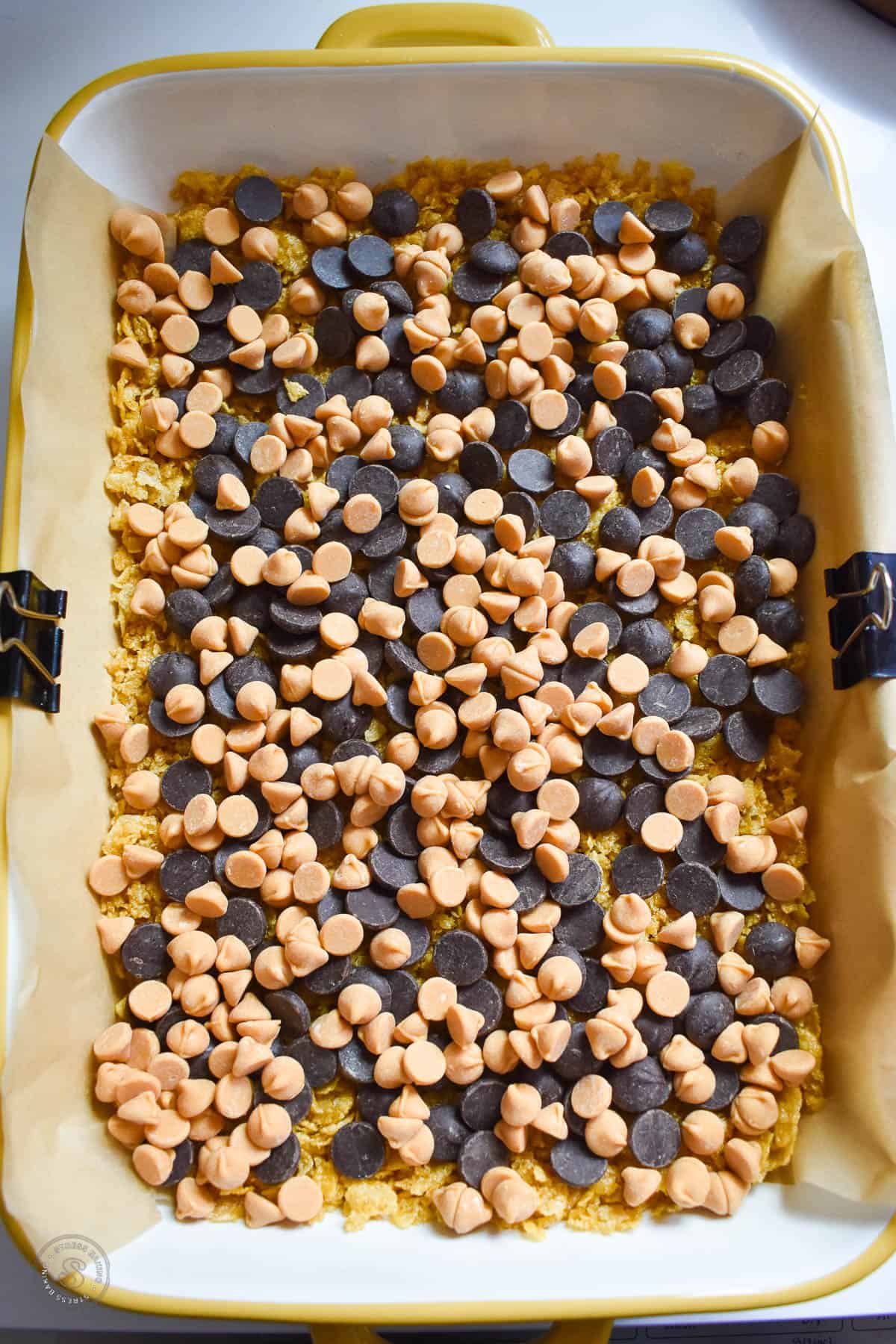 Chocolate chips and butterscotch chips scattered on top of rushed cornflake cereal pressed into a baking dish