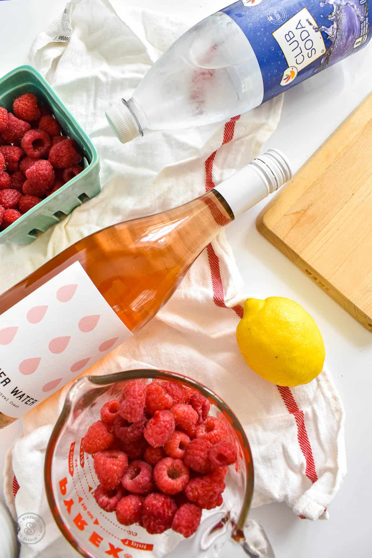 Ingredients for raspberry sangria on a white countertop including club soda, rose wine, raspberries and a lemon
