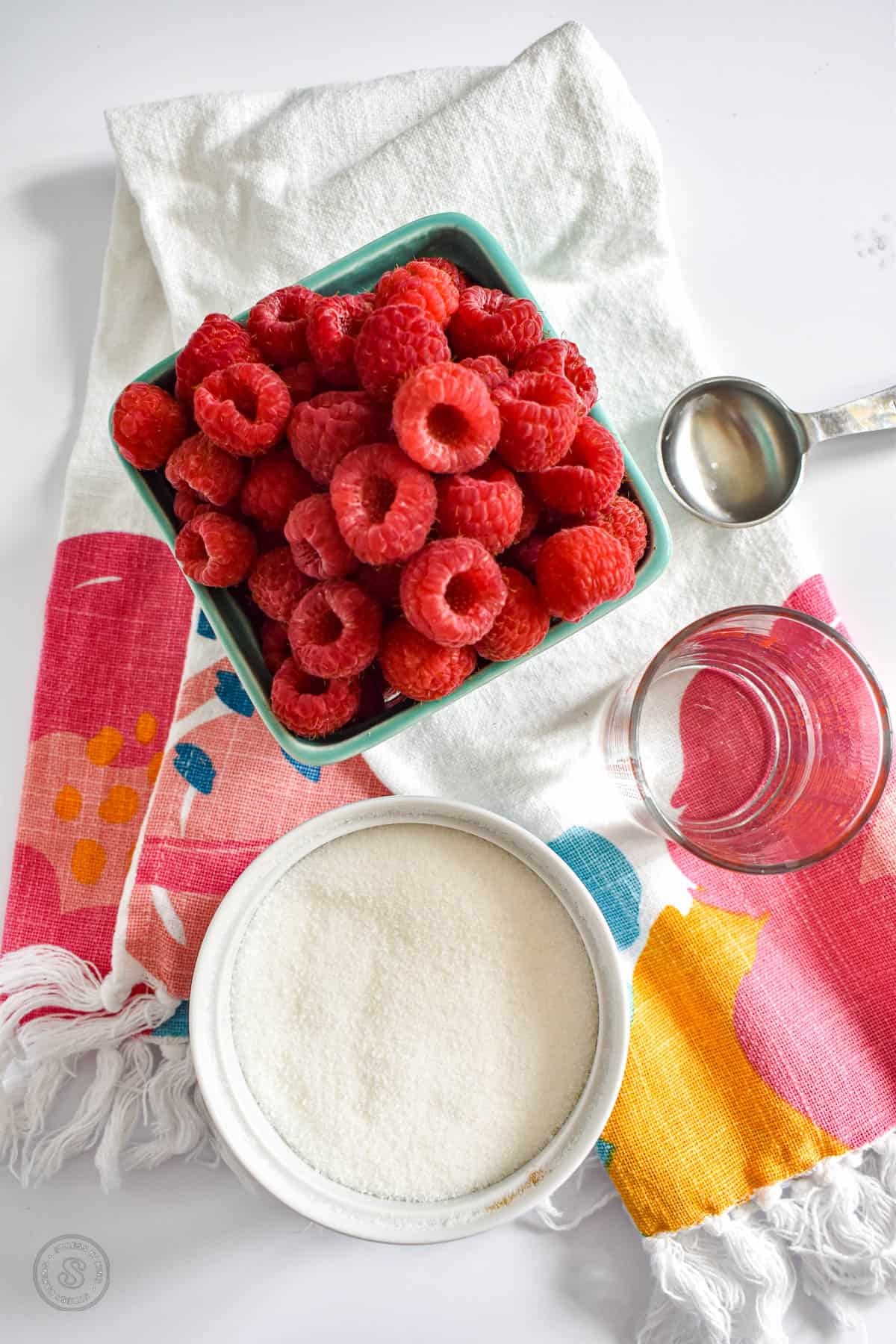 A teal container with bright red raspberries with a bowl of sugar and other utensils on a colorful dish towel