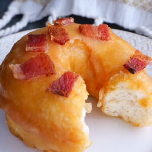 A maple donut topped with crisp bacon pieces with a bite taken out to display the soft fluffy inside