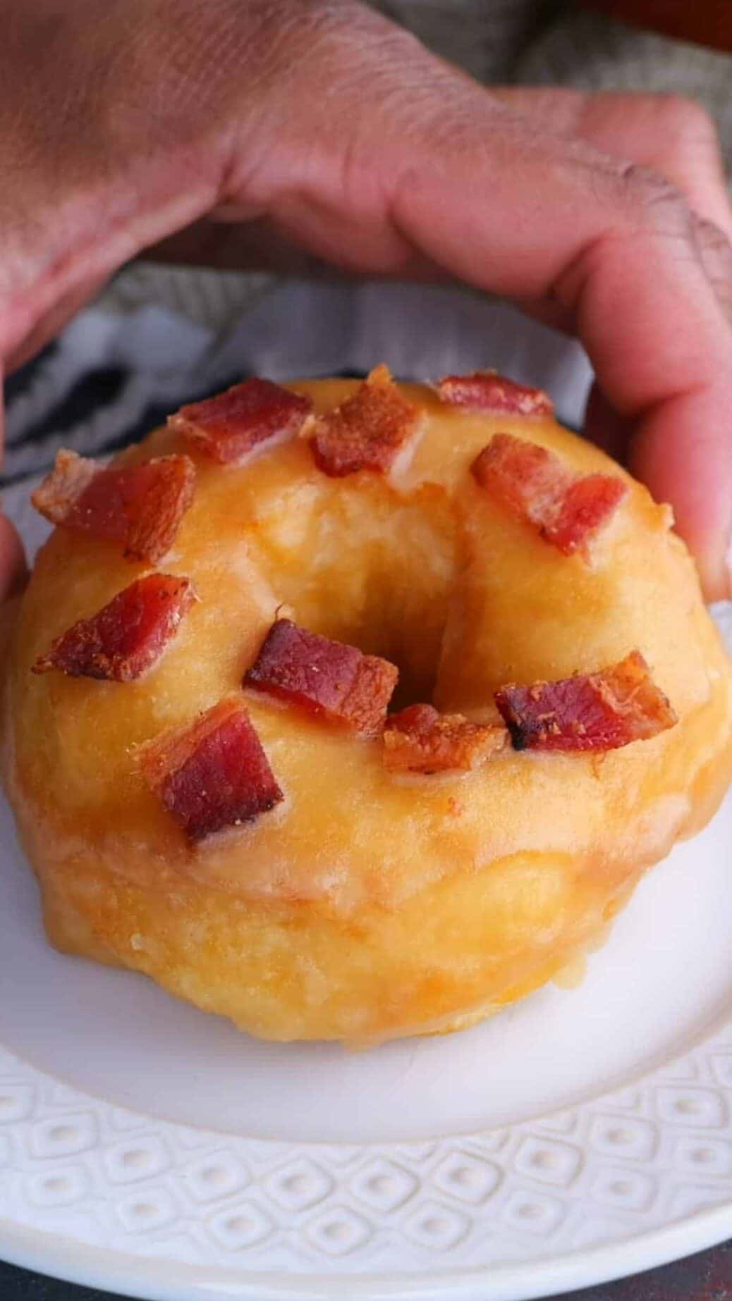 A maple donut topped with crisp bacon pieces being placed on a white plate