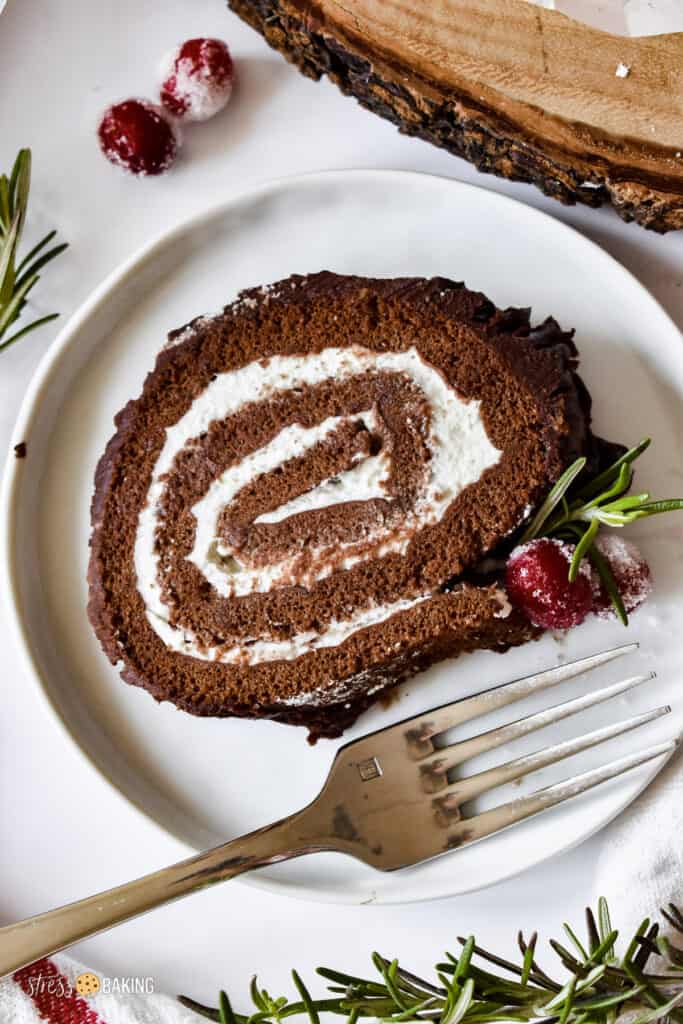 A slice of chocolate yule log cake filled with whipped cream and decorated with sugared cranberries and rosemary twigs