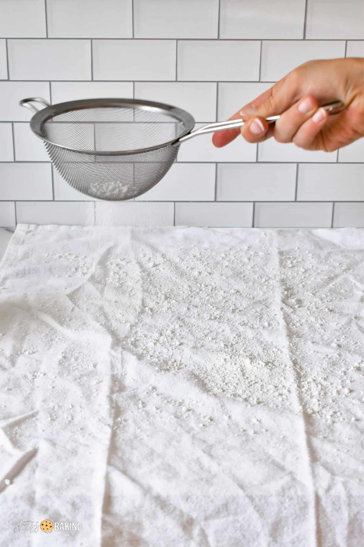 Using a fine mesh sieve to dust a white kitchen towel with powdered sugar