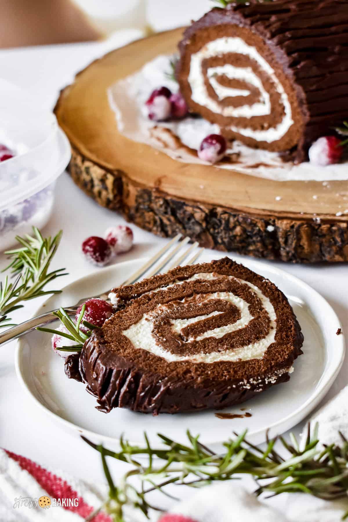 A slice of chocolate yule log cake filled with whipped cream and decorated with sugared cranberries and rosemary twigs